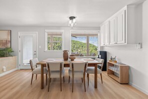A dining area with a wooden table and chairs, a vase, and a view of greenery through the windows. - Lakeview Nook - 6 min to Cypress Creek Park (Austin)