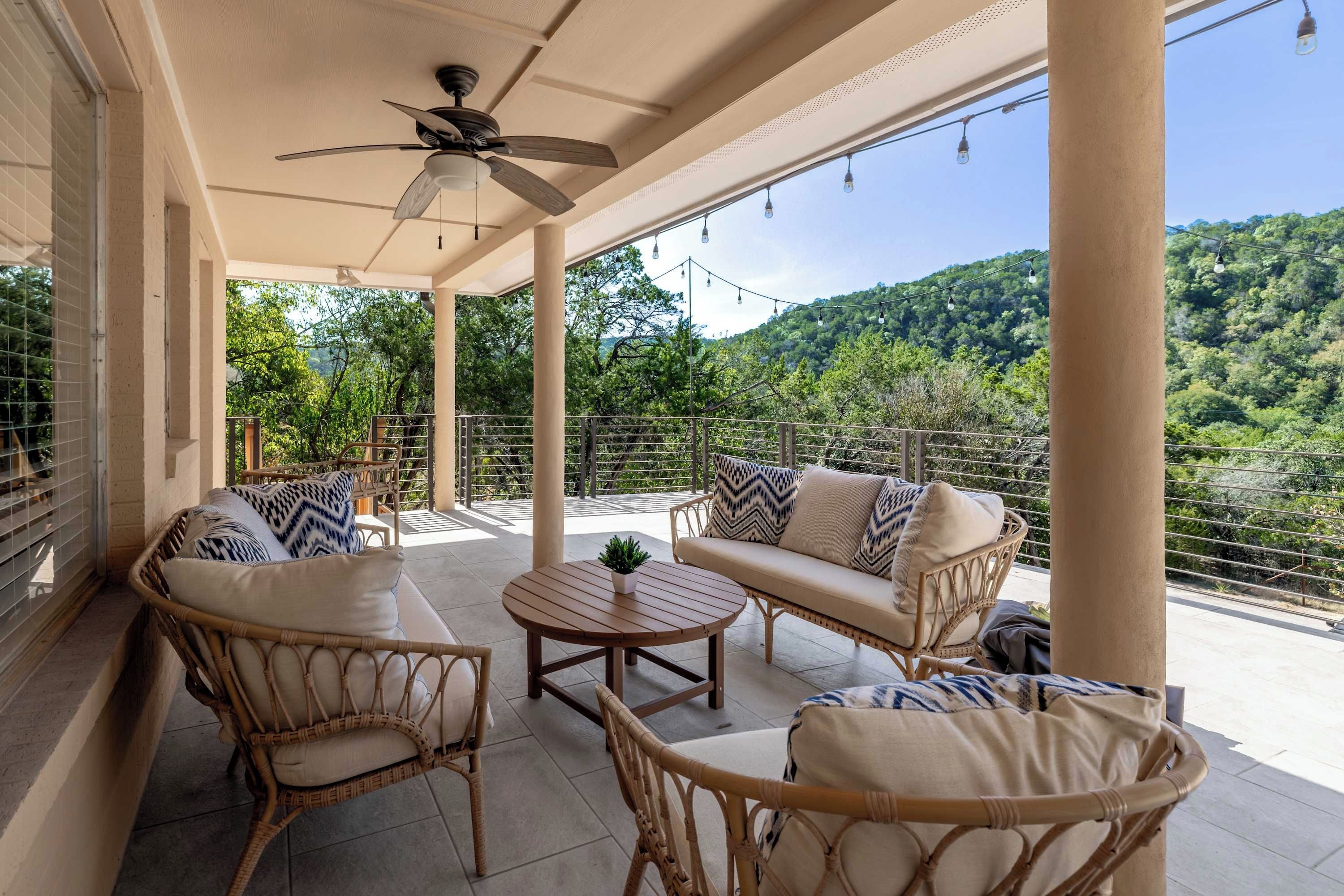 A covered outdoor seating area with wicker furniture, a ceiling fan, and a view of a forested hillside.