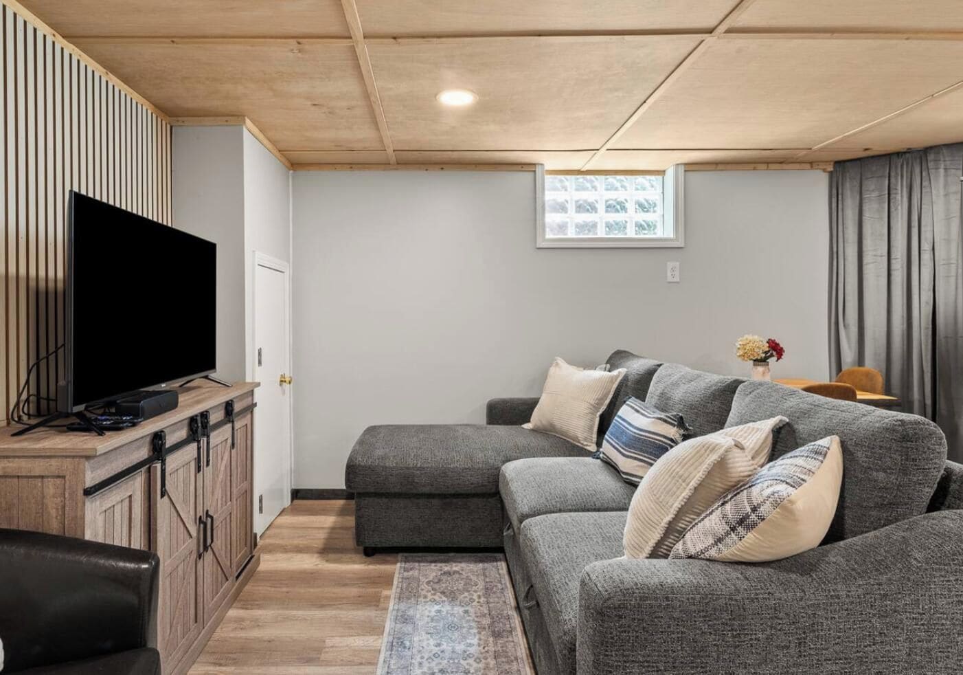 A modern living room with a flat-screen TV, grey sofa, wooden cabinets, and a small table with a vase.