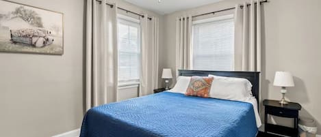 Une chambre à coucher avec un lit, deux tables de chevet, un ventilateur de plafond et une fenêtre avec des rideaux.