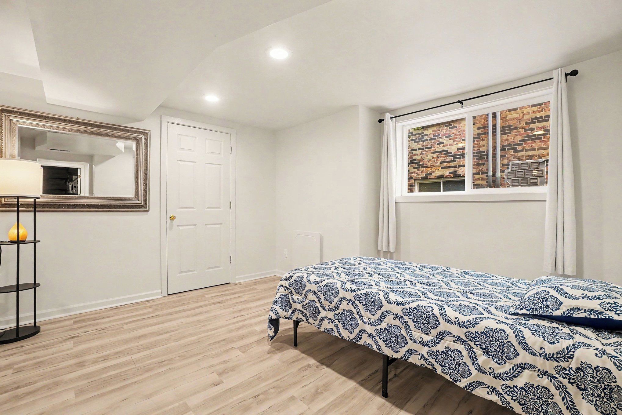 Ein Schlafzimmer mit Bett, Kleiderschrank, Spiegel und Fenster mit Blick auf eine Ziegelwand.