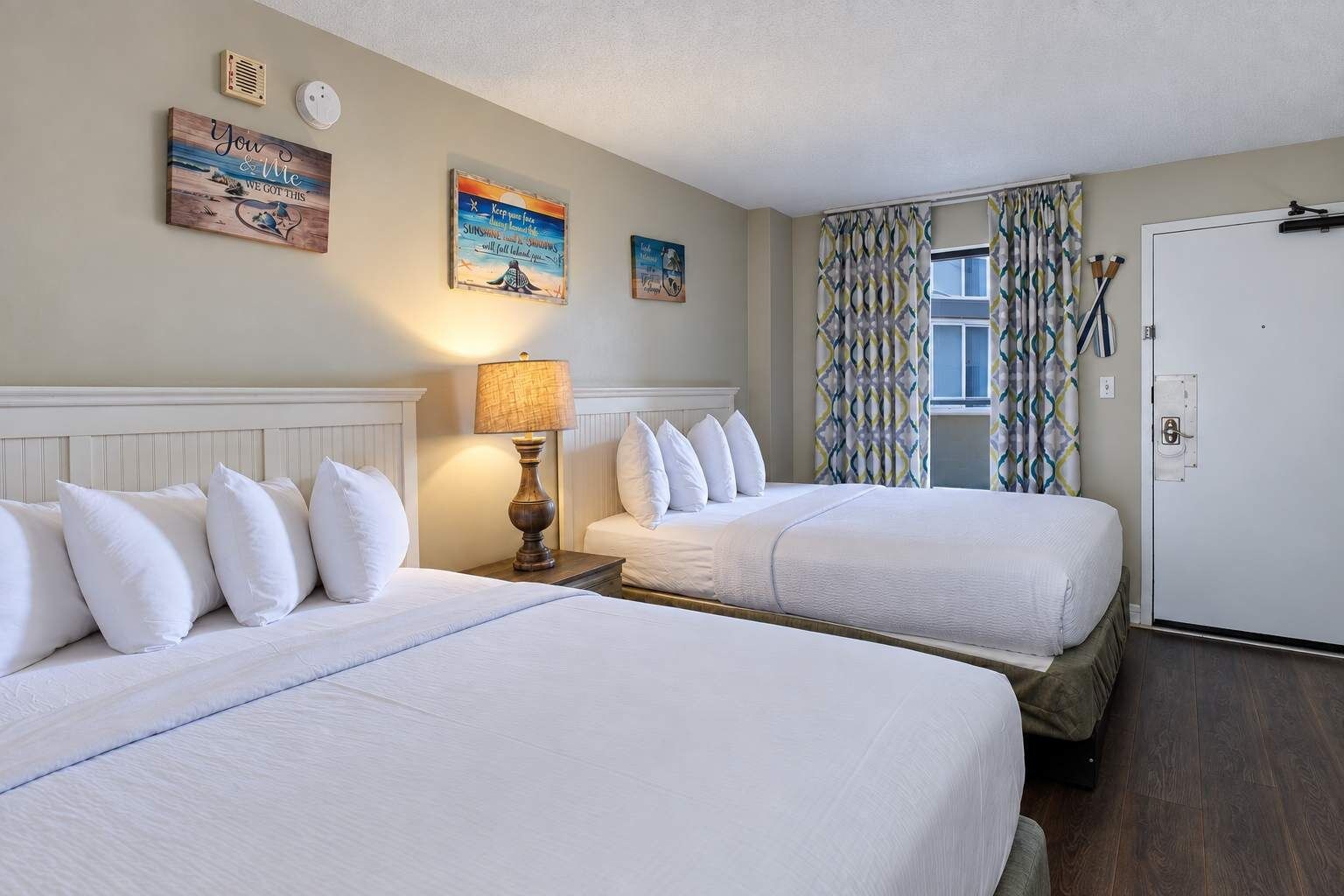 Een hotelkamer met twee bedden, een nachtkastje met een lamp, een raam met gordijnen en een kunstwerk aan de muur.