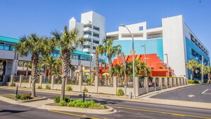 A modern hotel with a white facade, multiple floors, and a parking area with palm trees. - Cozy Oceanfront Getaway! Landmark 640 for 6! (Myrtle Beach)