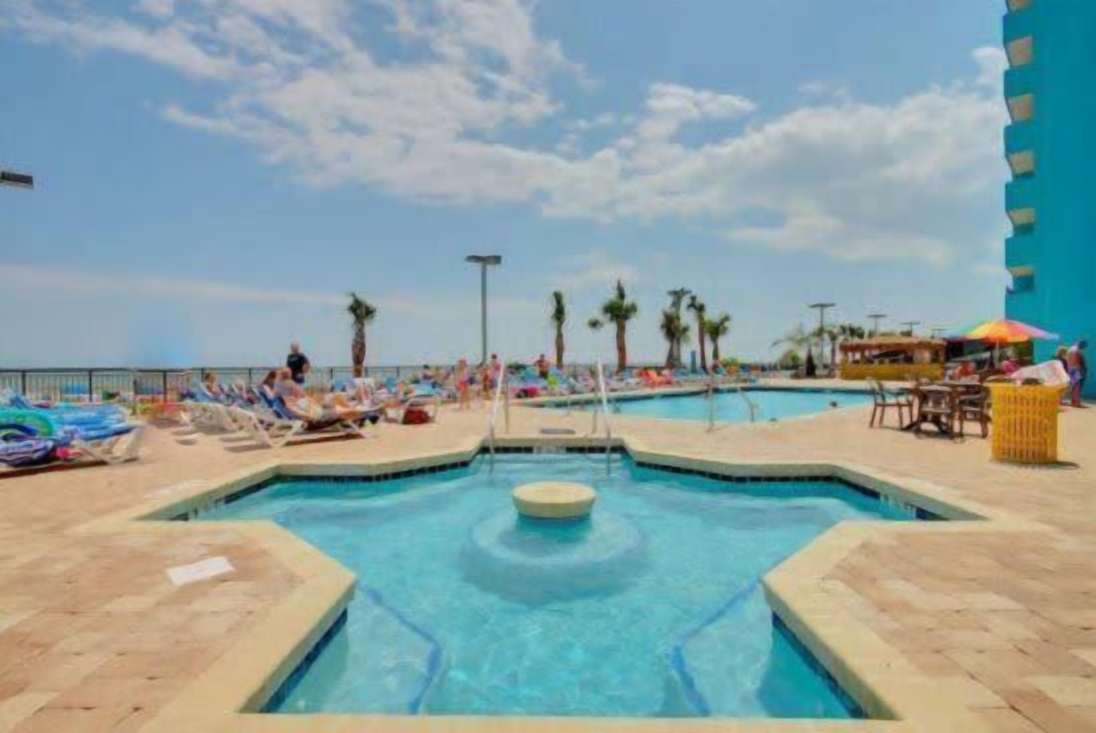 A pool area with lounge chairs and umbrellas, surrounded by palm trees and a clear sky.