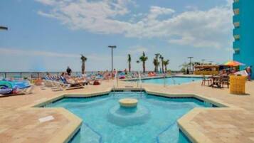 A pool area with lounge chairs and umbrellas, surrounded by palm trees and a clear sky.