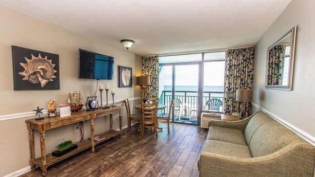 A living room with a wooden floor, a sofa, a dining table with chairs, and a balcony with a view. - Cozy Oceanfront Getaway! Landmark 640 for 6! (Myrtle Beach)