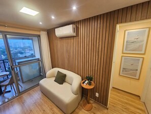 A modern living room with a sofa, a small round table with a plant, and a wall-mounted air conditioner. - Apartamento em Moema com varanda e vista da cidade (São Paulo)