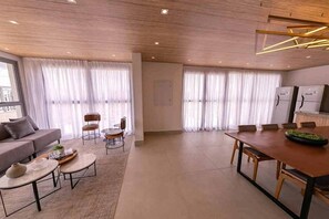 A modern living room with a sofa, coffee table, and chairs, a dining area with a table and chairs, and large windows with sheer curtains. - Apartamento em Moema com varanda e vista da cidade (São Paulo)