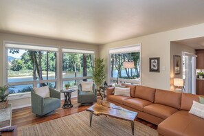 A spacious living room with a leather sofa, two armchairs, a coffee table, and large windows offering a view of a lake and trees.
