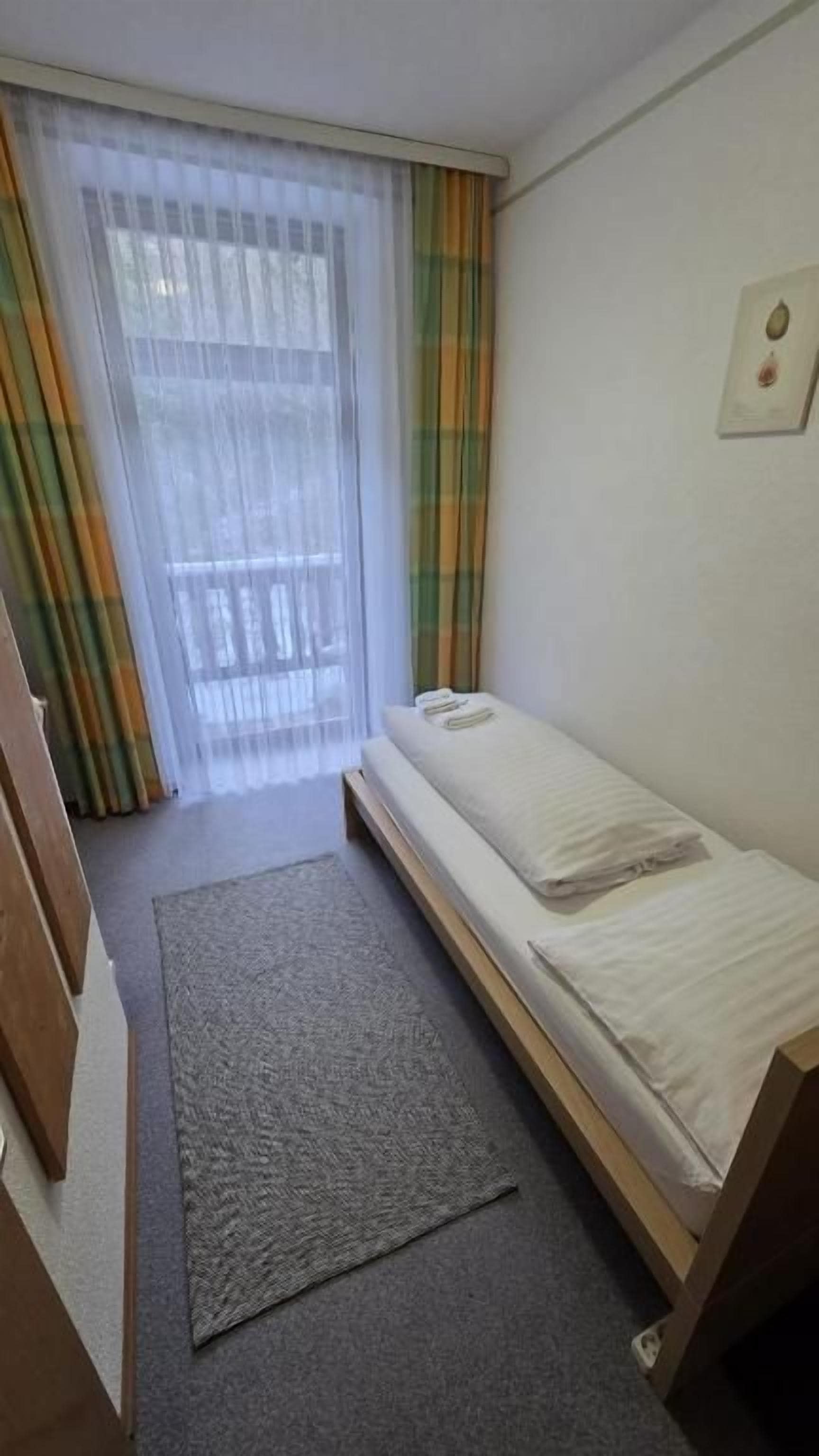 Una habitación pequeña con una cama individual, una ventana con cortinas traslúcidas, una alfombra pequeña y un cuadro en la pared.