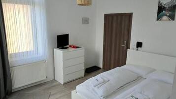Una camera d'albergo con un letto, una scrivania, una sedia, un comò, una televisione e una finestra con tende.