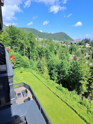 A view from a balcony overlooking a well-maintained lawn with a wooden deck, dense greenery, and a distant village.