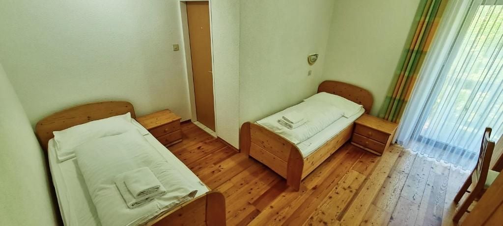 Une pièce avec deux lits en bois, une table de chevet en bois, une porte et une fenêtre avec des rideaux légers.