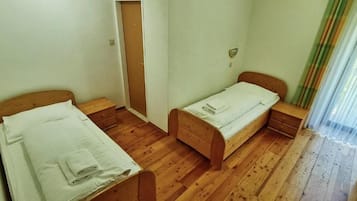 Une pièce avec deux lits en bois, une table de chevet en bois, une porte et une fenêtre avec des rideaux légers.