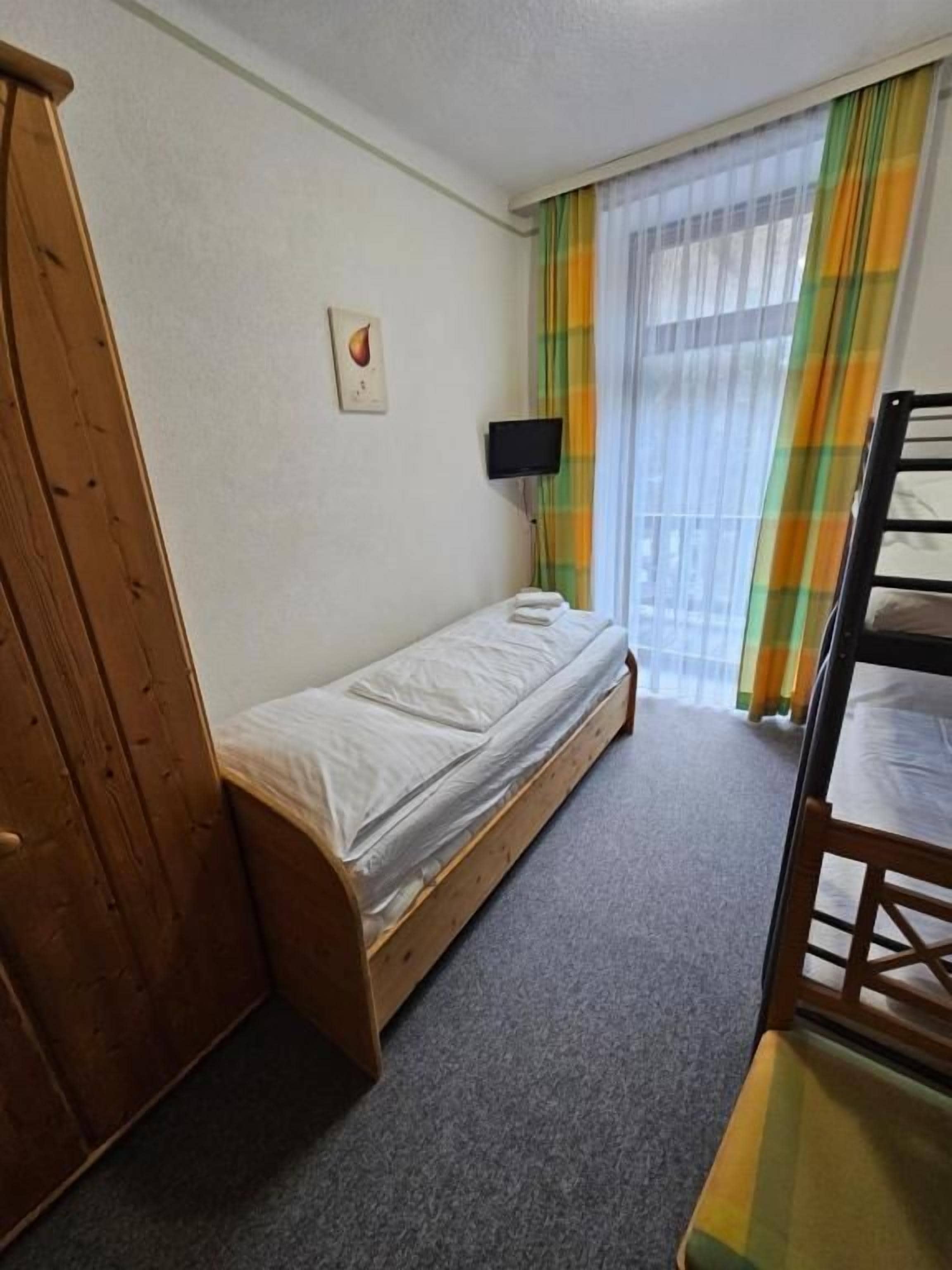 Ein Einzelbett mit Holzrahmen, ein kleiner Nachttisch, ein an der Wand montierter Fernseher und ein Fenster mit Vorhängen.