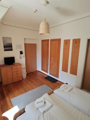 A bedroom with a bed, wooden wardrobe, a desk with a chair, a hanging light fixture, and a door leading to another room.