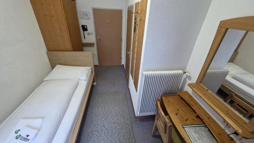 A small, compact room with two single beds, a wooden bench, a radiator, and a door leading to another room.