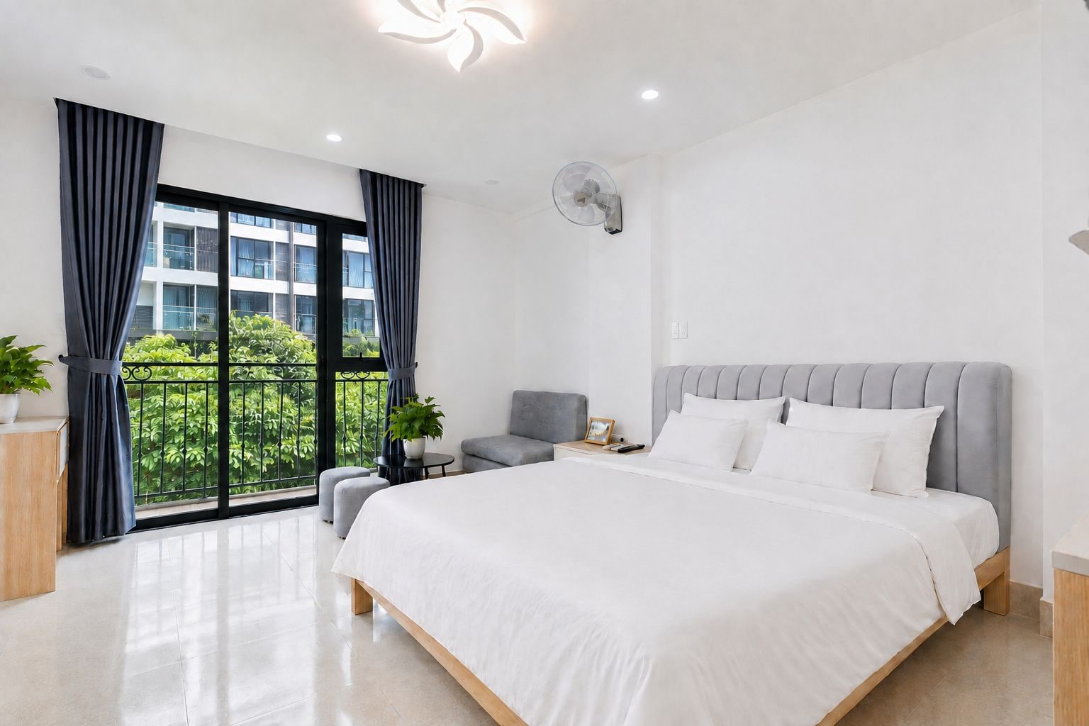 A modern bedroom with a large bed, a grey upholstered chair, a small table with a lamp, and a view of greenery through a balcony.