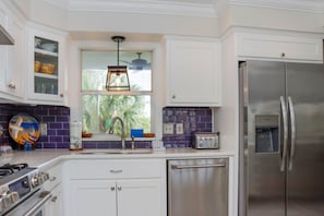 A modern kitchen with white cabinets, a stainless steel refrigerator, a built-in oven, a window with a view of palm trees, and a hanging pendant light. - Boardwalk Bungalow (Port St. Joe)