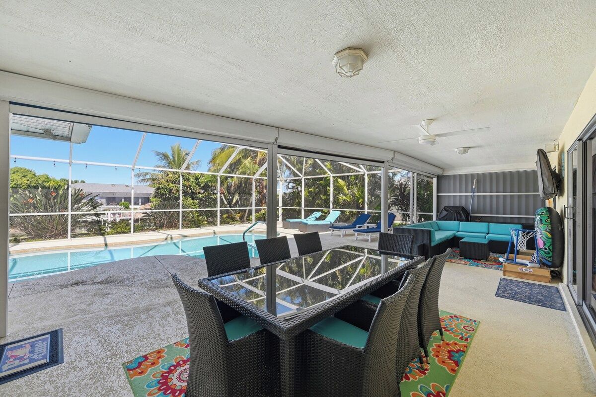 Área externa coberta com mesa de vidro, cadeiras, sofá e piscina ao fundo.