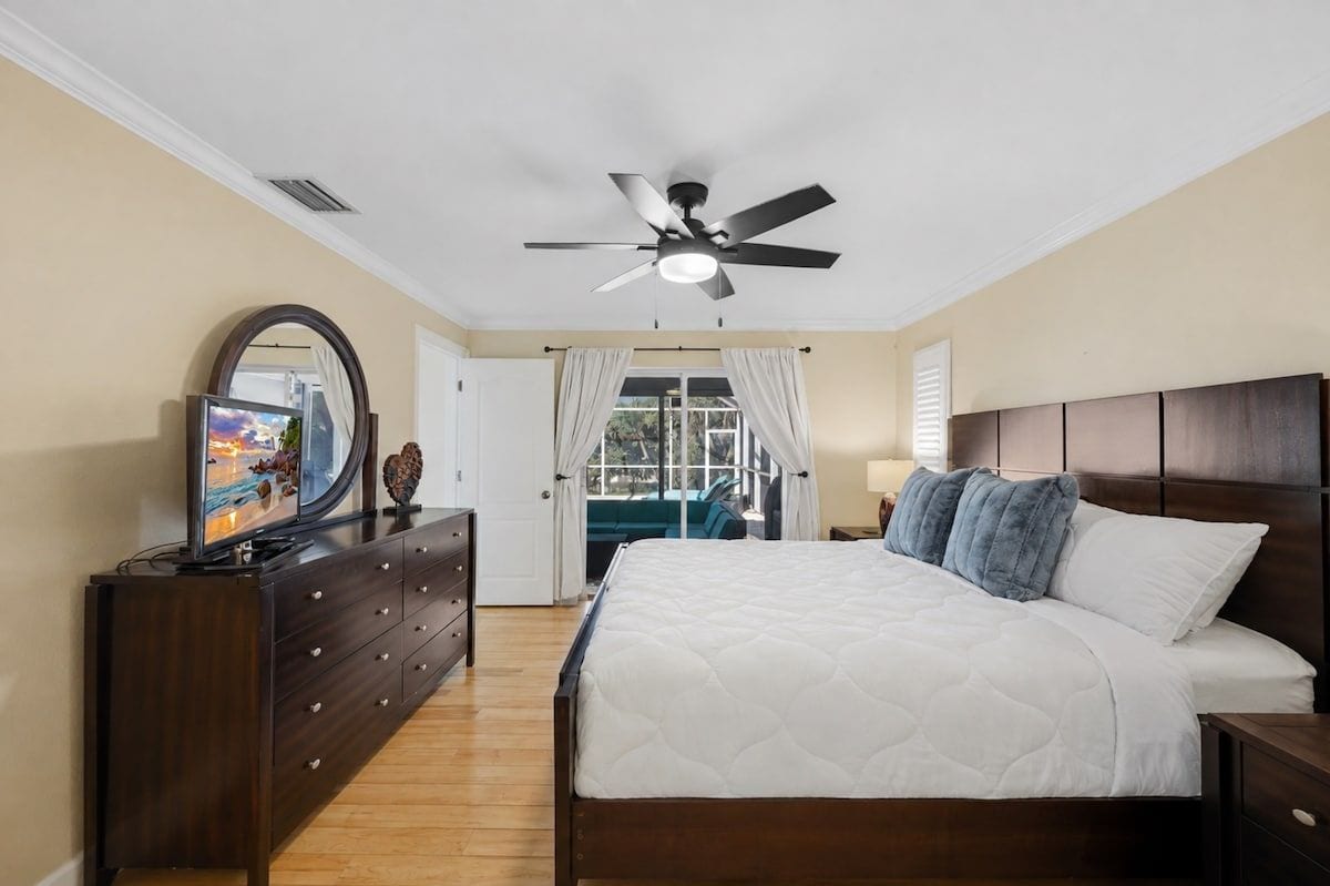 A bedroom with a bed, a dresser, a ceiling fan, a TV on the dresser, and a window with curtains.