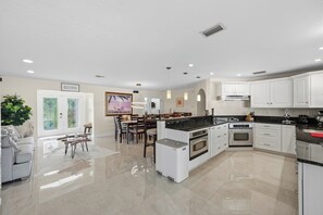 A modern kitchen with white cabinets, a central island, and a dining area with a white sofa and chairs. - Piece of Paradise | 3BR Waterfront Pool Retreat (Cape Coral)