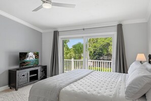 A bedroom with a bed, a television on a dresser, a balcony with a view of greenery, and a ceiling fan. - The Palms at McGregor | Luxury 4BR Pool Home (Fort Myers)