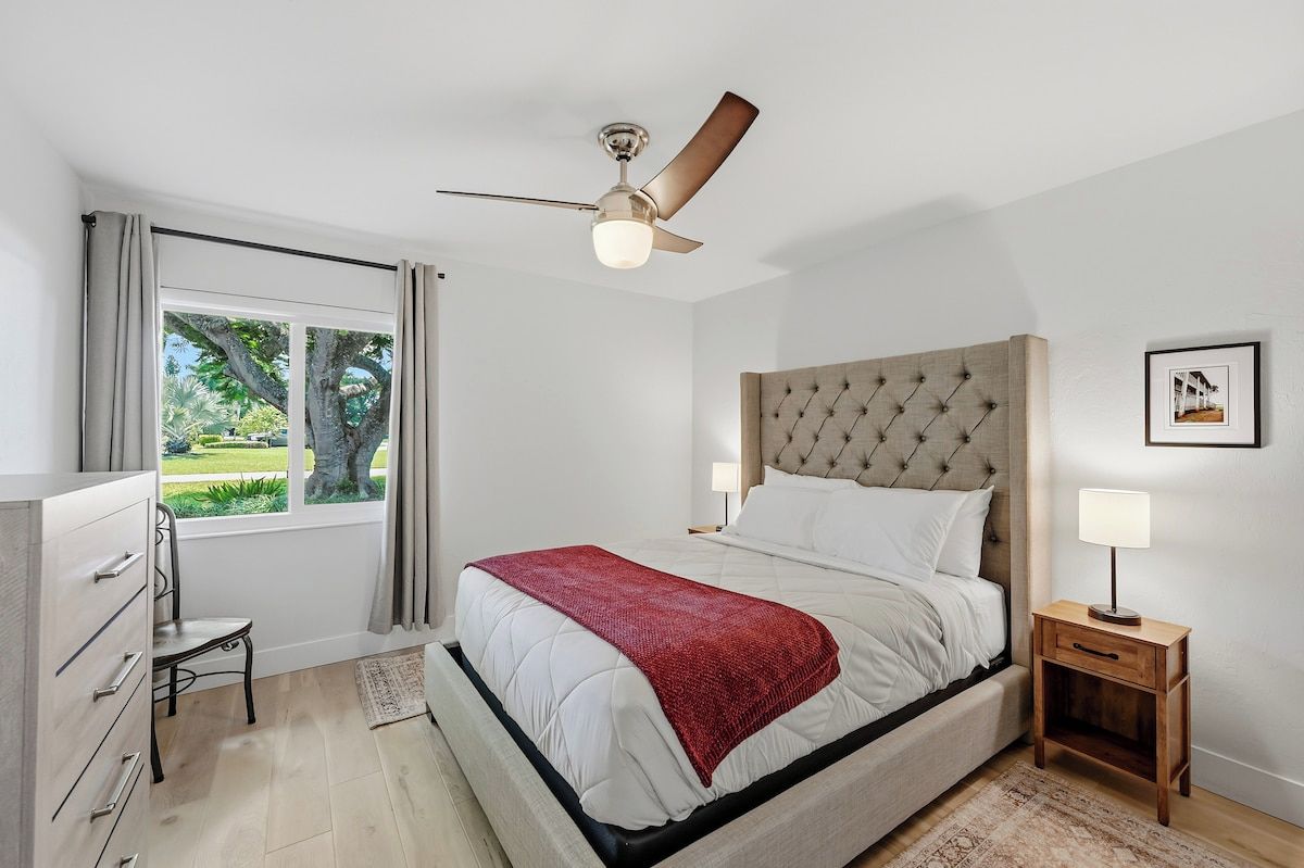 A bedroom with a bed, a ceiling fan, a bedside table with a lamp, a window with curtains, and a view of the outdoors.