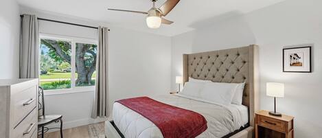 Un dormitorio con cama, ventilador de techo, mesita de noche con lámpara, ventana con cortinas y vistas al exterior.