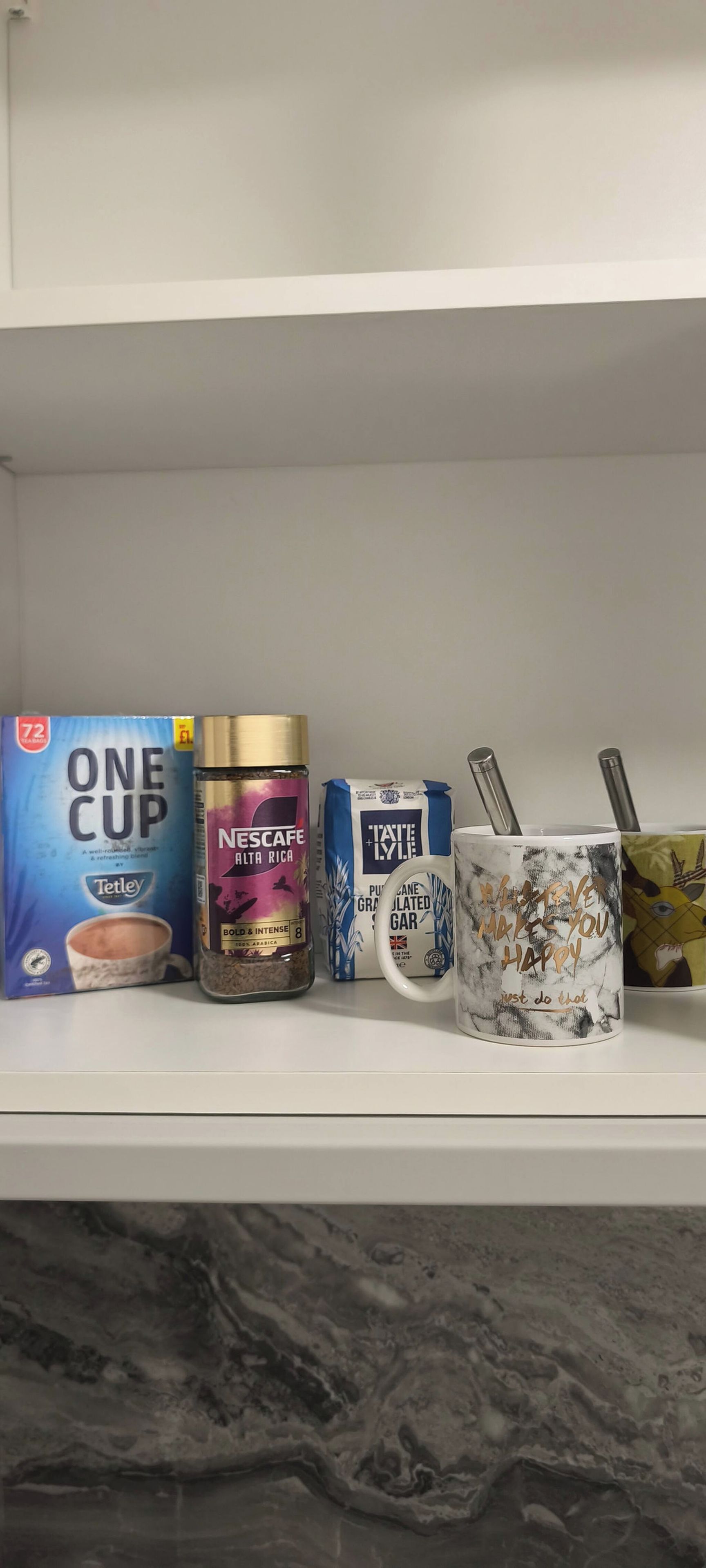 A shelf with a box of Tetley One Cup tea, a jar of Nescafe Malta Rica coffee, a pack of Tate & Lyle sugar, and a coffee mug with a motivational quote.