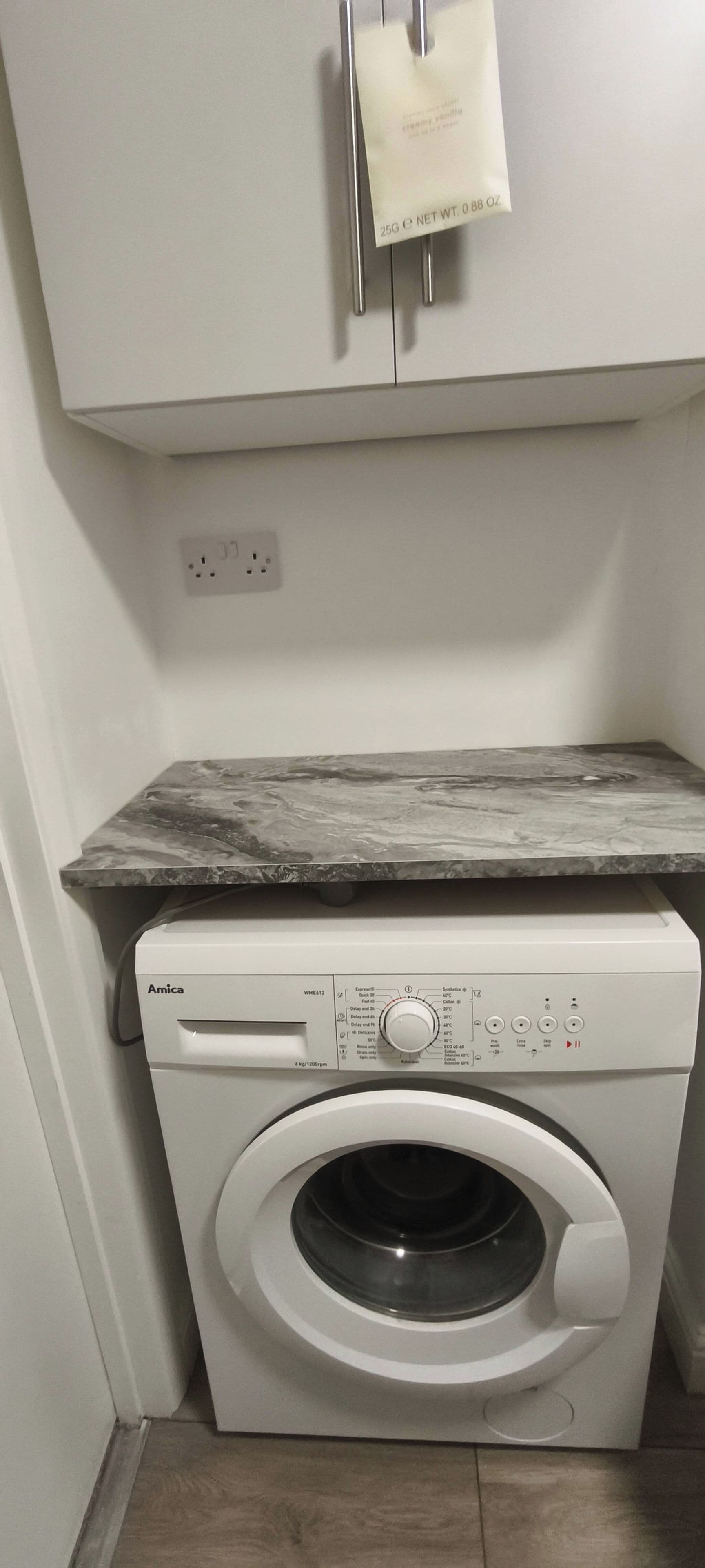 Una lavadora Amica blanca y compacta con un panel de control, una encimera de mármol encima y un armario blanco con dispensador de jabón.