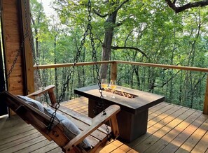 A wooden deck with a swing chair, a table with a fire pit, and a view of a forest. - Tree Loft: Your romantic treetop escape near Hot Springs. (Bismarck)