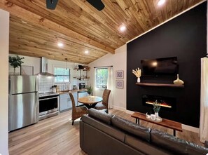 A modern kitchen with stainless steel appliances, a dining area with a round table and chairs, and a living space with a black wall-mounted TV and fireplace. - Tree Loft: Your romantic treetop escape near Hot Springs. (Bismarck)