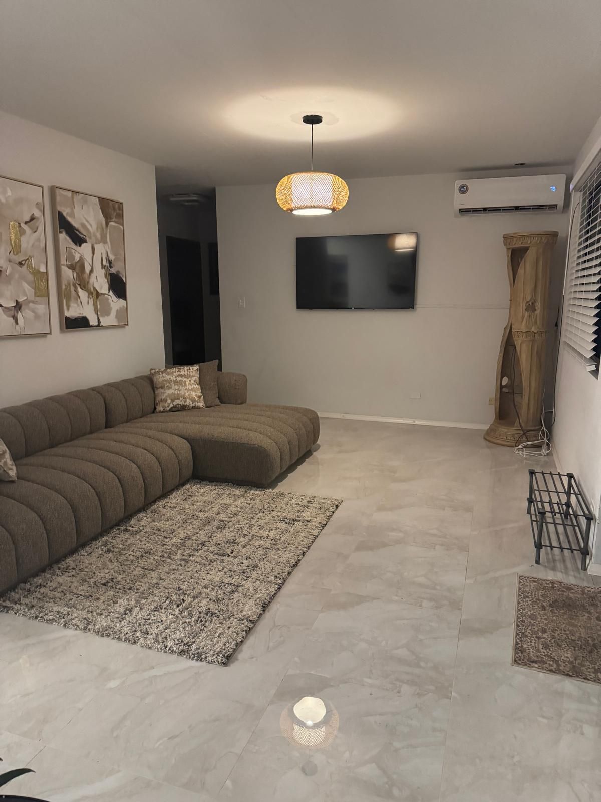 Un salon moderne avec un canapé d’angle, un téléviseur à écran plat, un luminaire suspendu et un vase décoratif.