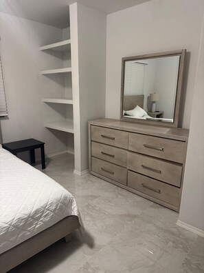 A bedroom with a bed, a dresser with drawers, a mirror, and a shelving unit.