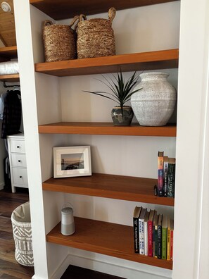 A bookshelf with woven baskets, a potted plant, a vase, a framed picture, and a row of books. - Pacific Edge Retreat · Surf, Trails & A Beautiful Home · Jordan River BC (Juan De Fuca (Part 2))