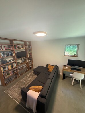 A living room with a dark grey sofa, a wooden bookshelf, a desk with a computer, and a window with a view of greenery. - Pacific Edge Retreat · Surf, Trails & A Beautiful Home · Jordan River BC (Juan De Fuca (Part 2))