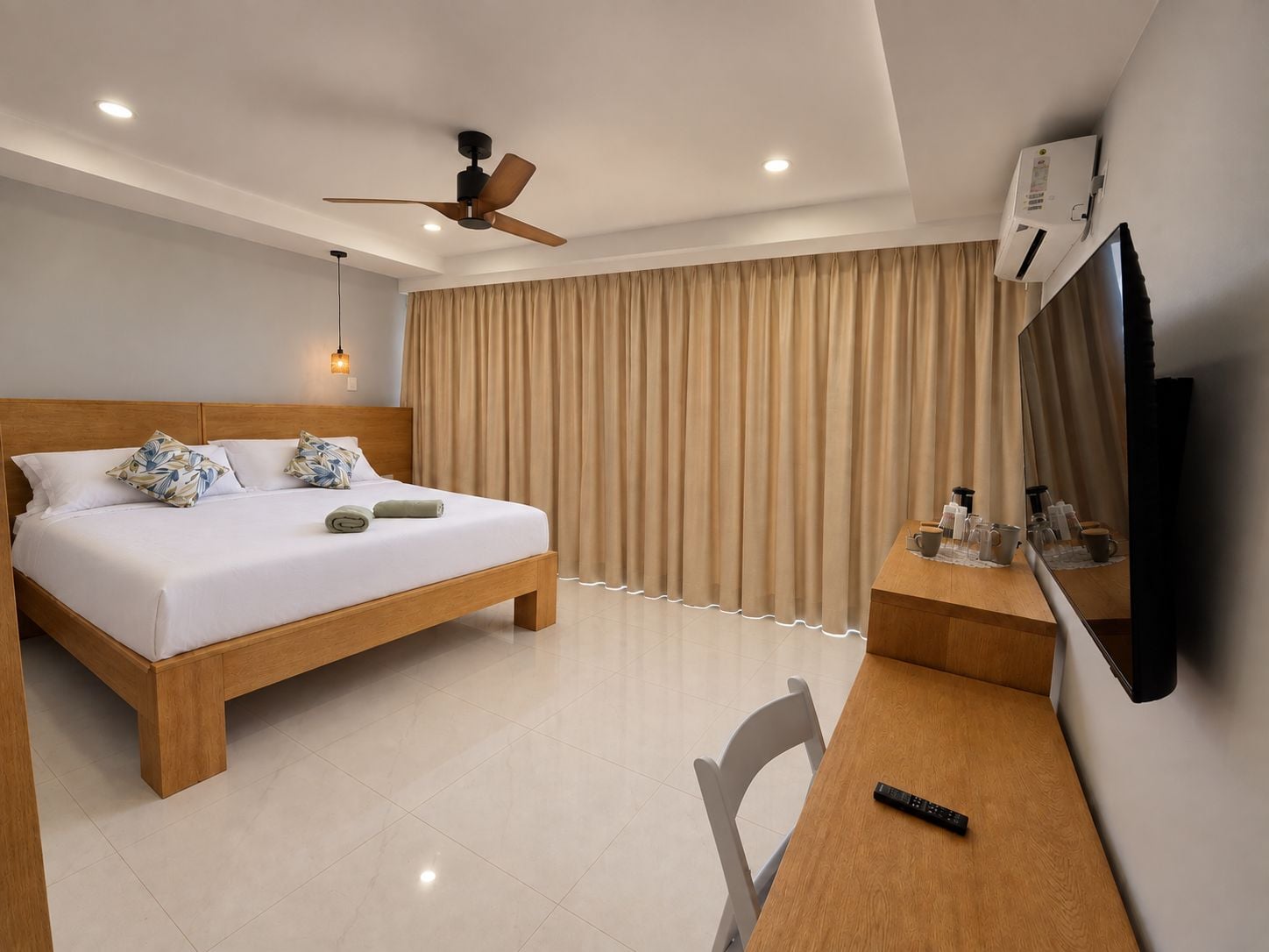 Ein Hotelzimmer mit einem Bett, einem Schreibtisch, einem Sessel, einem Deckenventilator, einem Fernseher und Vorhängen.
