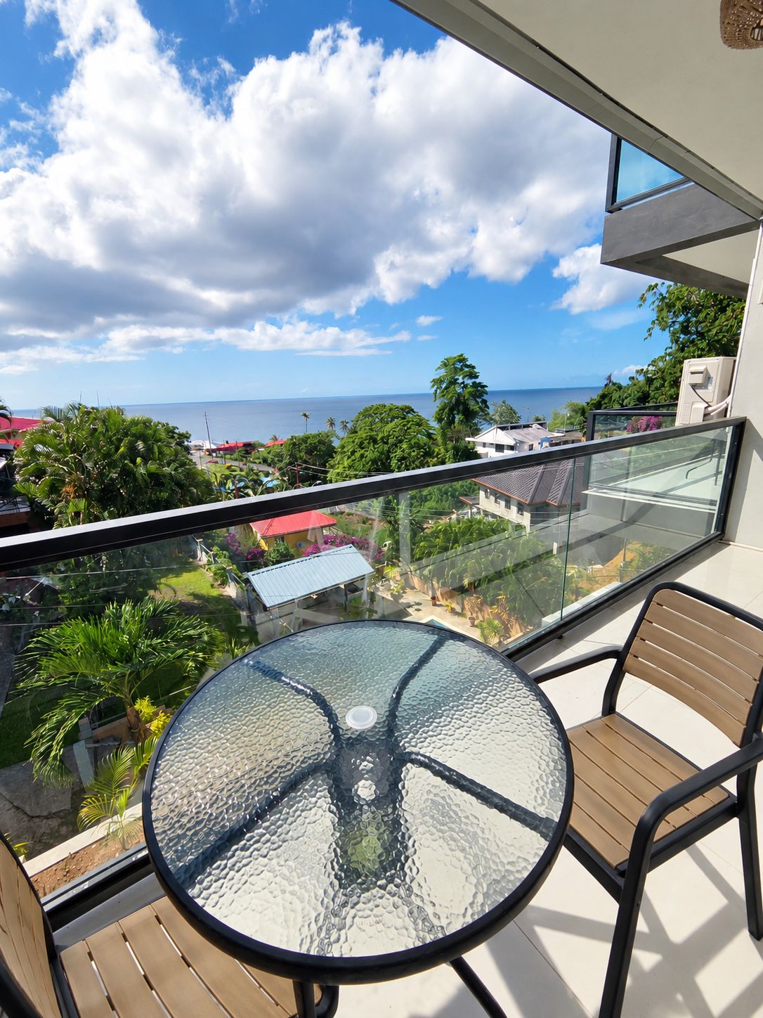A balcony with a glass table and chairs, overlooking a residential area and the sea.