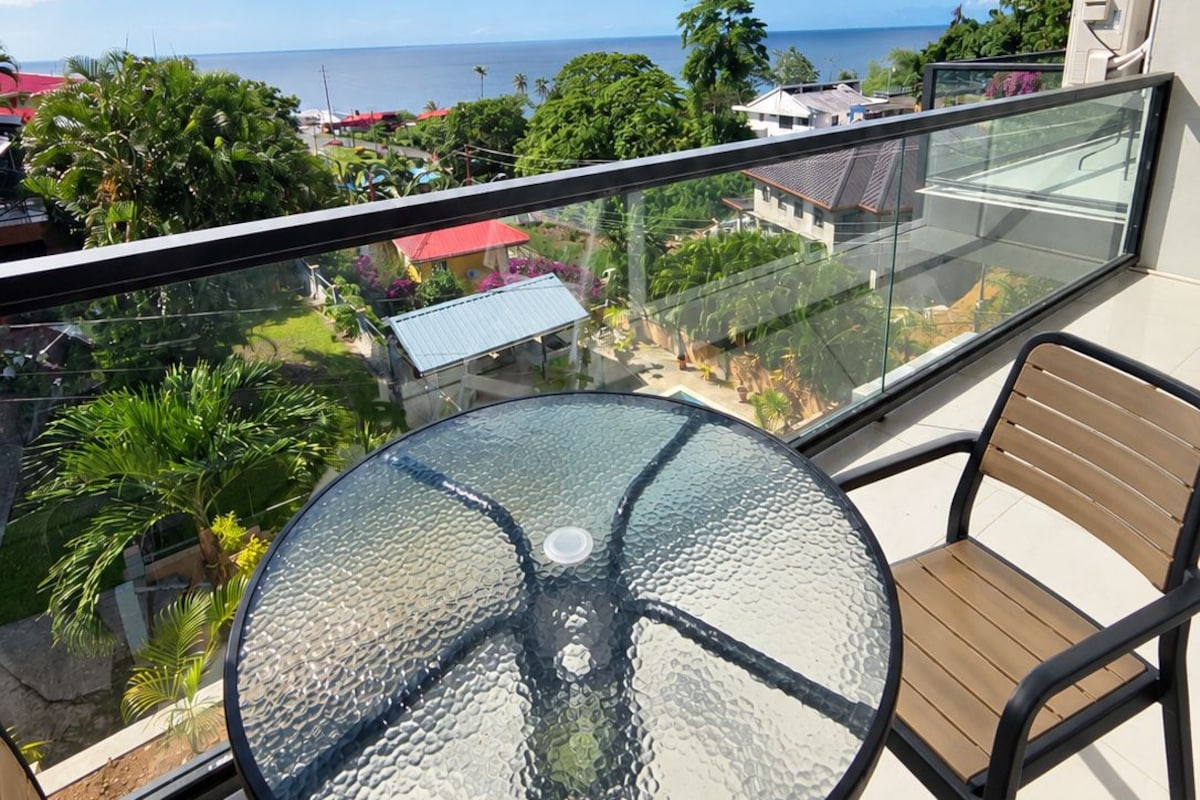 A balcony with a glass table and chairs, overlooking a residential area and the sea.