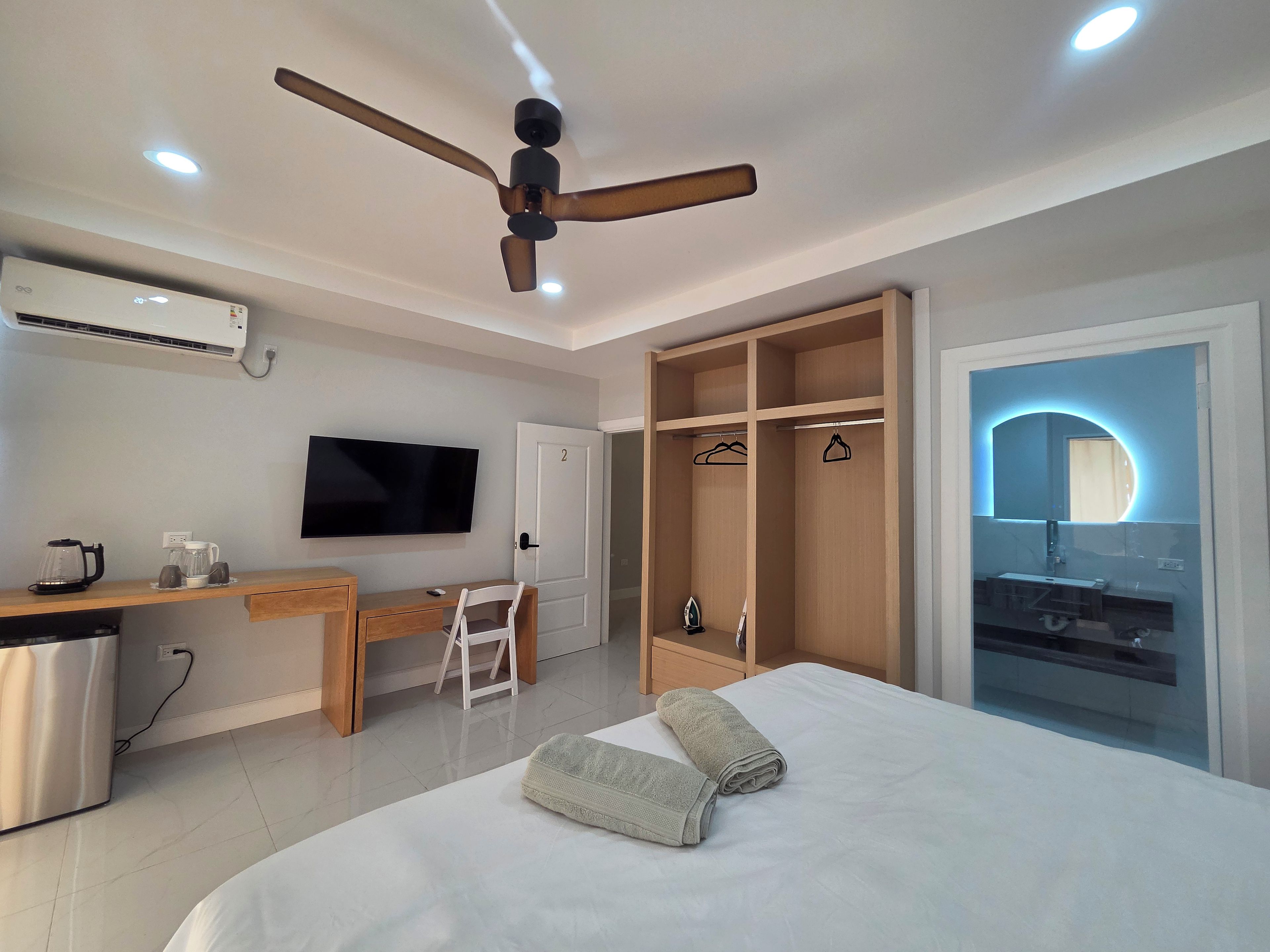Ein Hotelzimmer mit einem Bett, einem Schreibtisch mit Computer, einem Fernseher, einem Kleiderschrank und einem Deckenventilator.