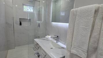 Un baño moderno con ducha envuelta en vidrio, un espejo grande y un tocador blanco con toallero.