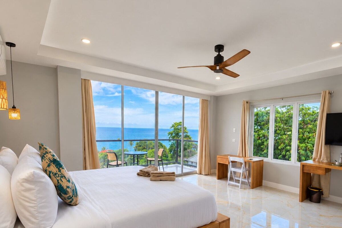A spacious bedroom with a large bed, a ceiling fan, a desk, and a view of the ocean.
