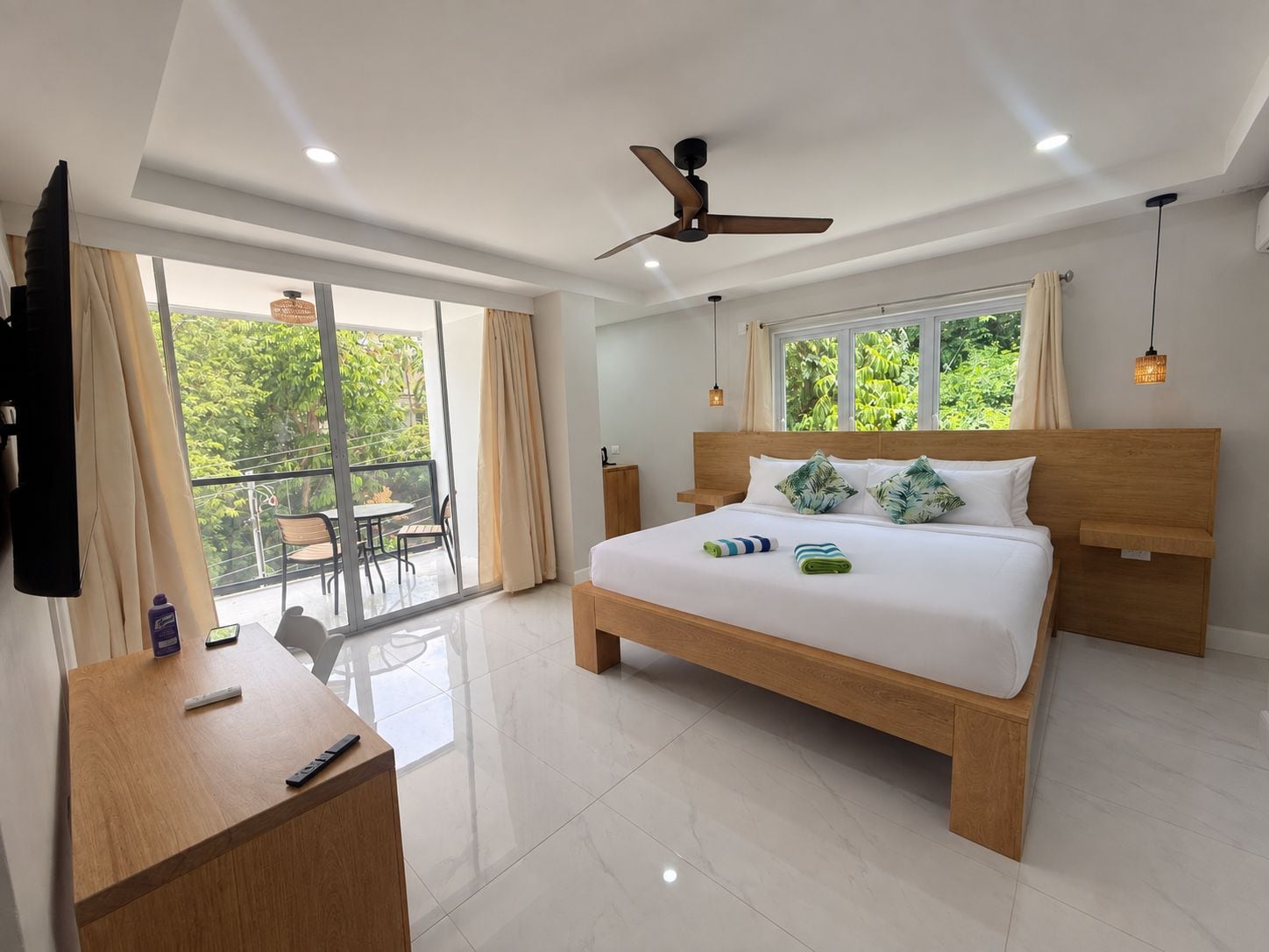 A modern hotel room with a large bed, a desk, a ceiling fan, and a balcony with a view of greenery.