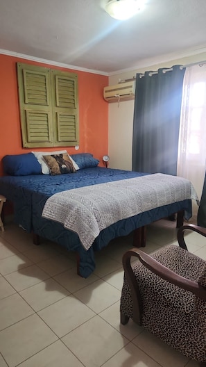 A bedroom with a bed, a chair, a window with curtains, an air conditioning unit, and a wall-mounted shelf.