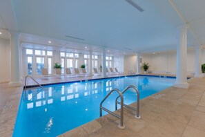 Une piscine intérieure à l’eau d’un bleu cristallin, entourée de colonnes blanches et baignée de lumière grâce à de grandes fenêtres.