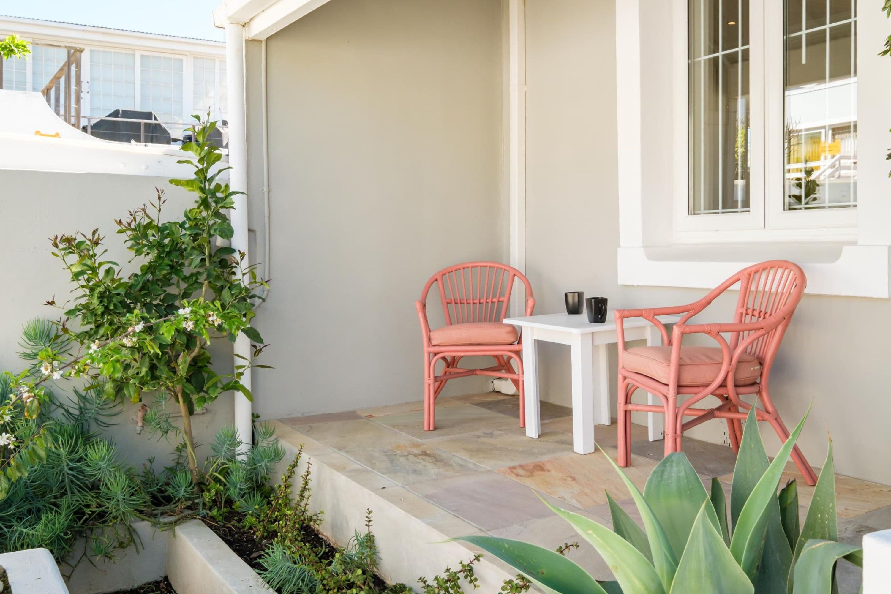 A small outdoor patio with two pink chairs and a white table, surrounded by various plants and a window.