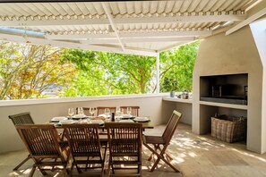 An outdoor dining area with a table set for four, surrounded by wooden chairs, under a white pergola with a view of trees. - Green Point 4 Bed Family Home w Pool Happy House (Cape Town)