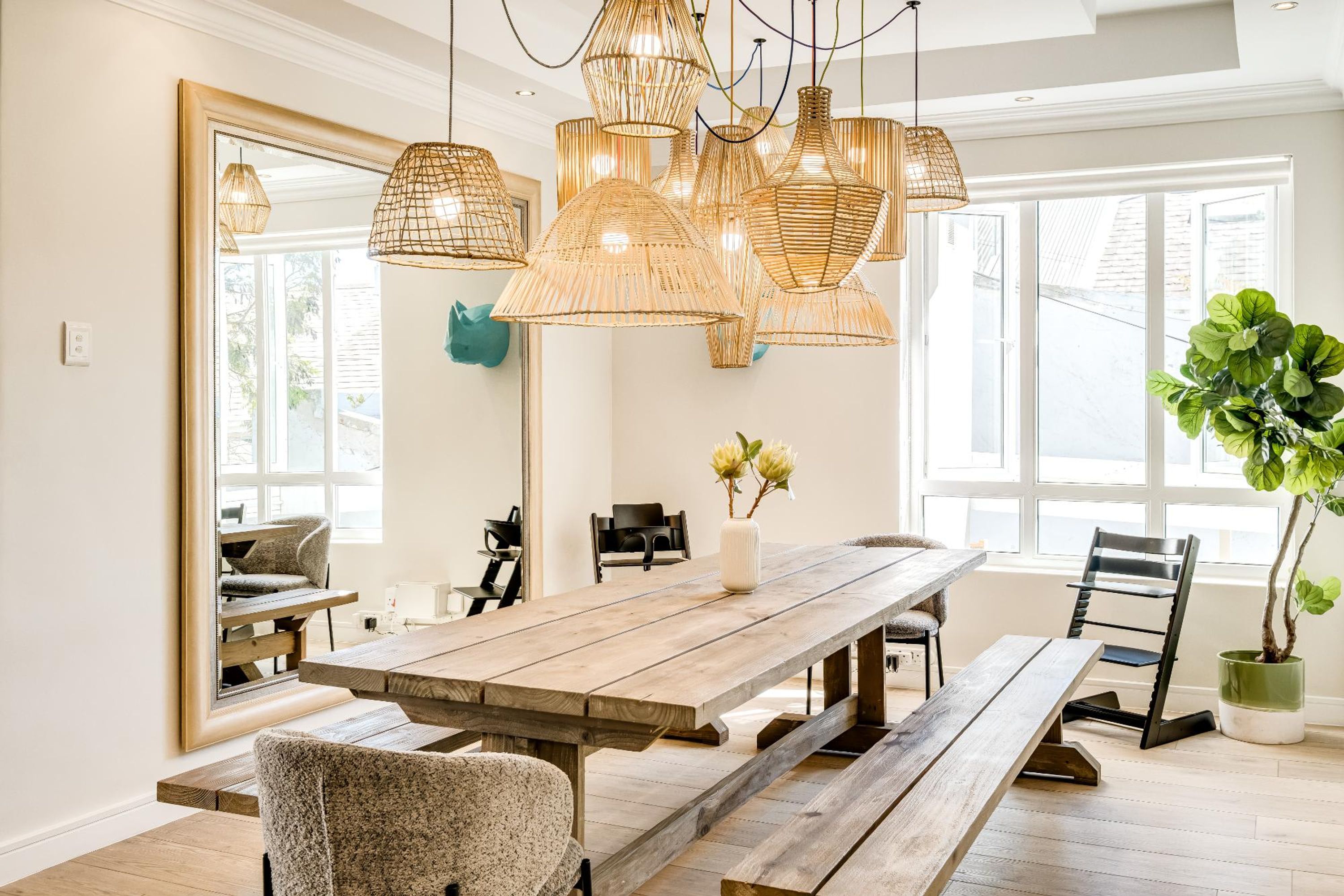 A dining area with a wooden table, benches, wicker pendant lights, and a large mirror on the wall.