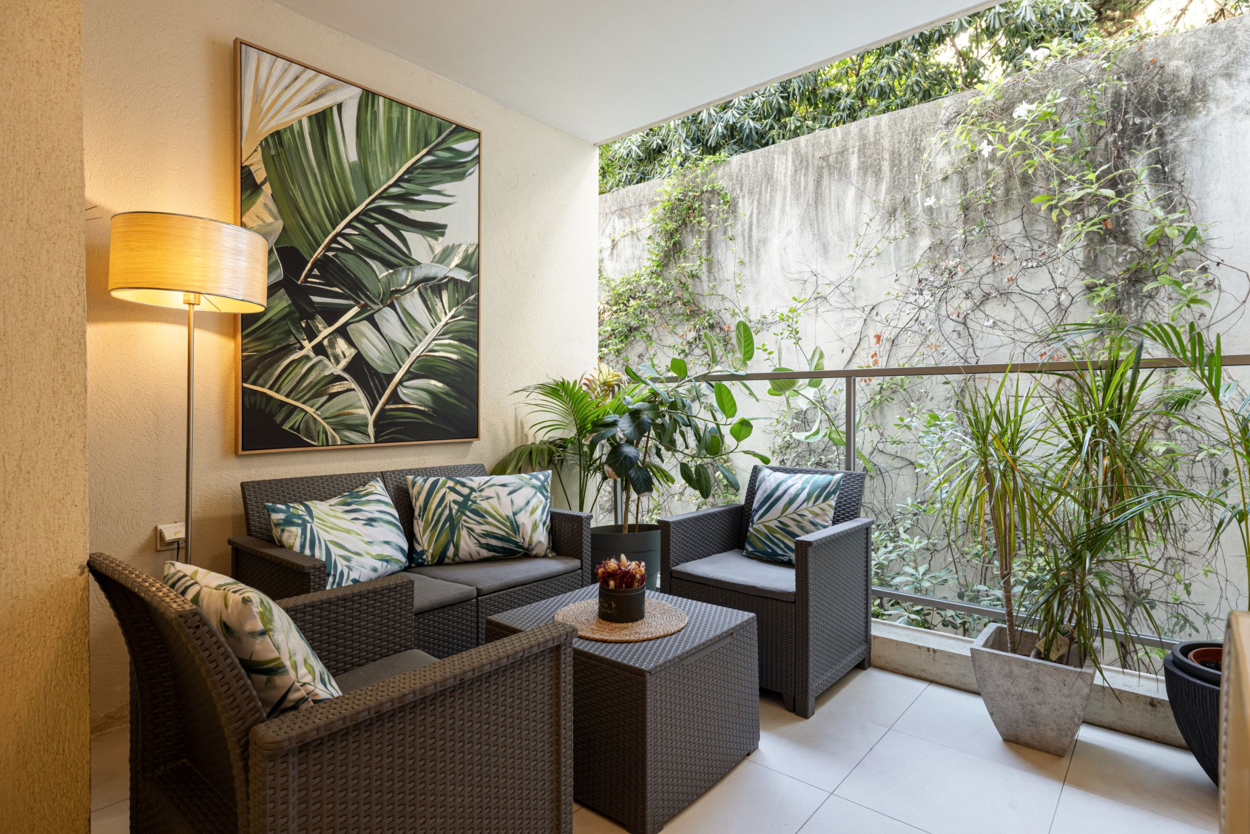 A modern outdoor seating area with wicker furniture, a wall-mounted lamp, and a large tropical leaf painting.